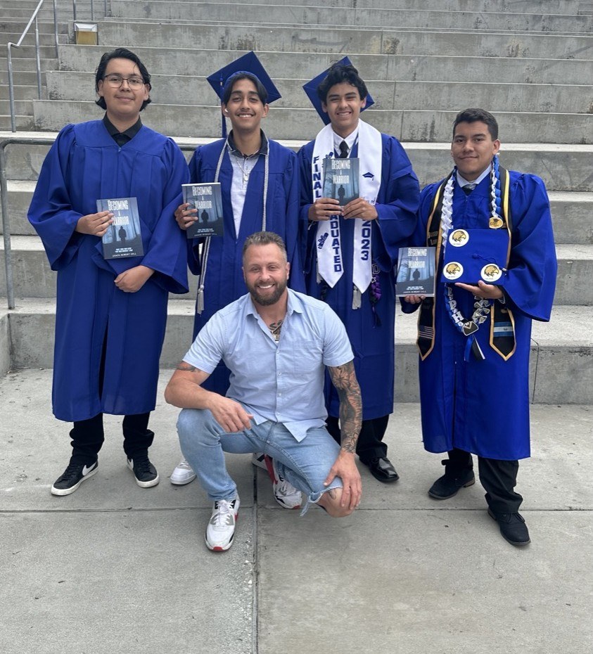 Young men graduating in cap and gown holding the Becoming a Warrior book with Jason Hill, Güerote
